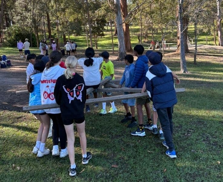 Year 6 students carrying a wodden stretcher over grass surrounded by trees