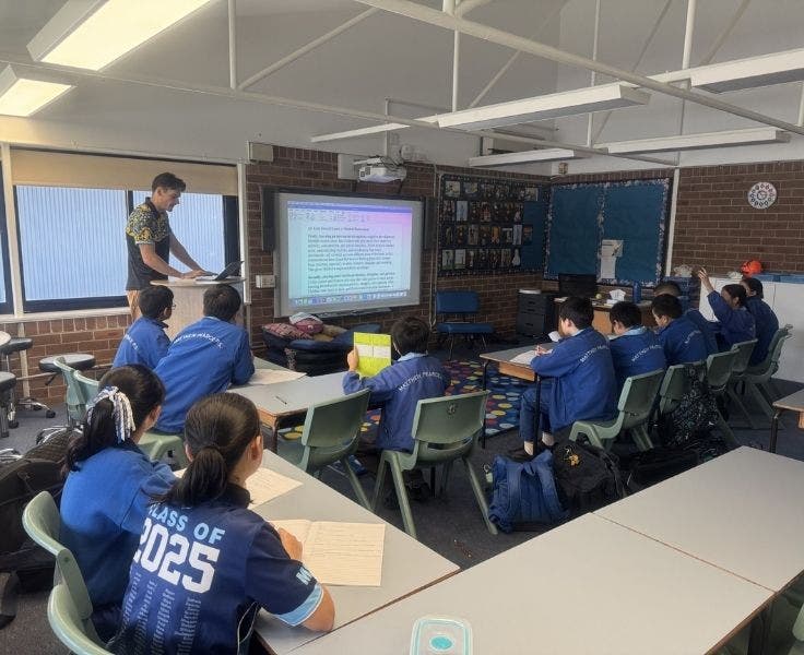 Students in a classroom taking notes with a teacher standing out the front
